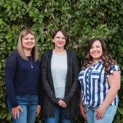 Three team members standing side by side outdoors in front of a lush green wall of foliage, wearing casual professional clothing and smiling toward the camera.
